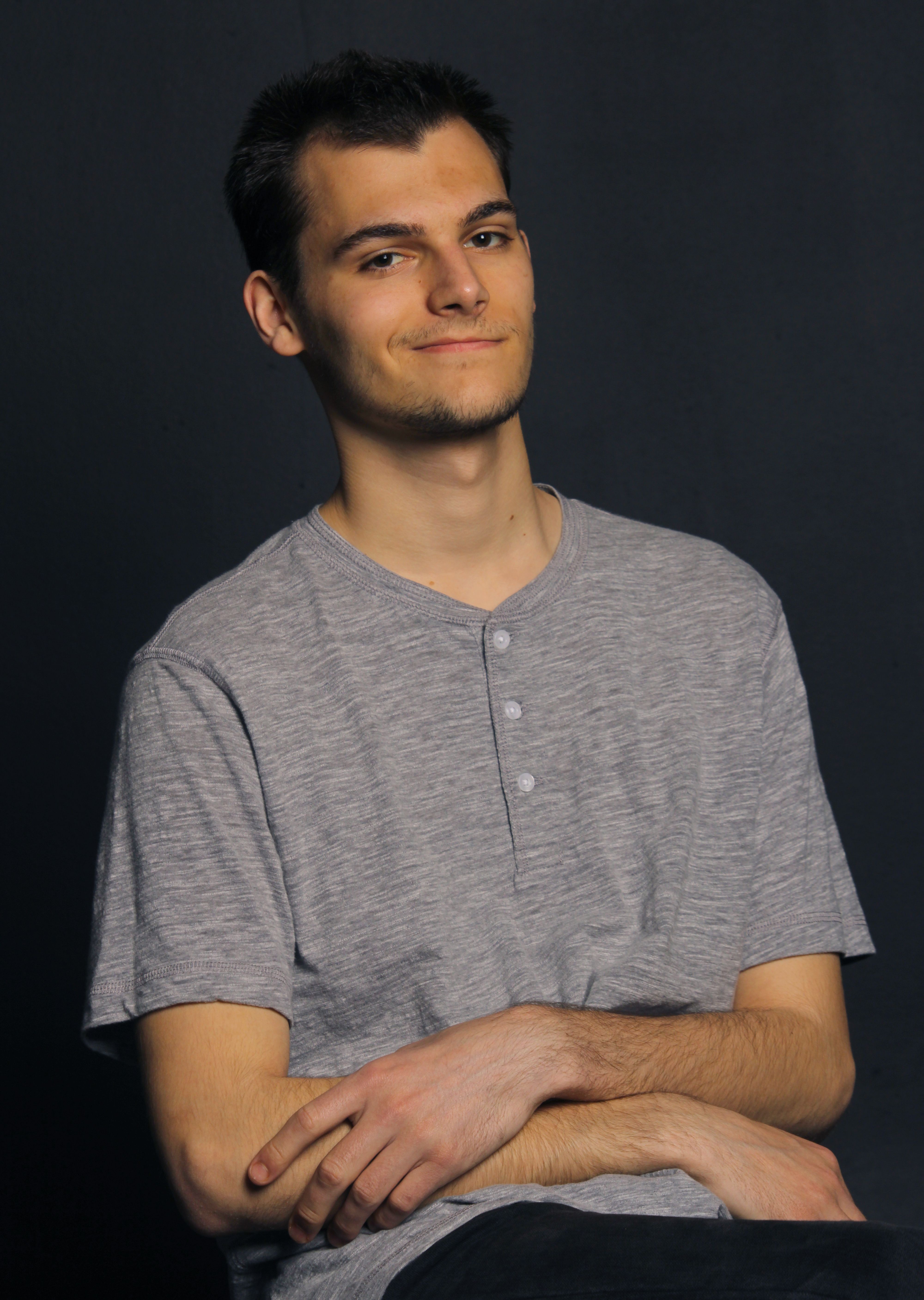 Portrait of a young man in a gray shirt with a dark background, exuding a relaxed and confident demeanor.