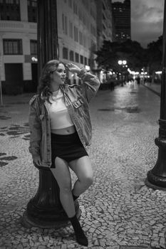 Stylish woman poses on a lit street in Curitiba at night, wearing a denim jacket and skirt.