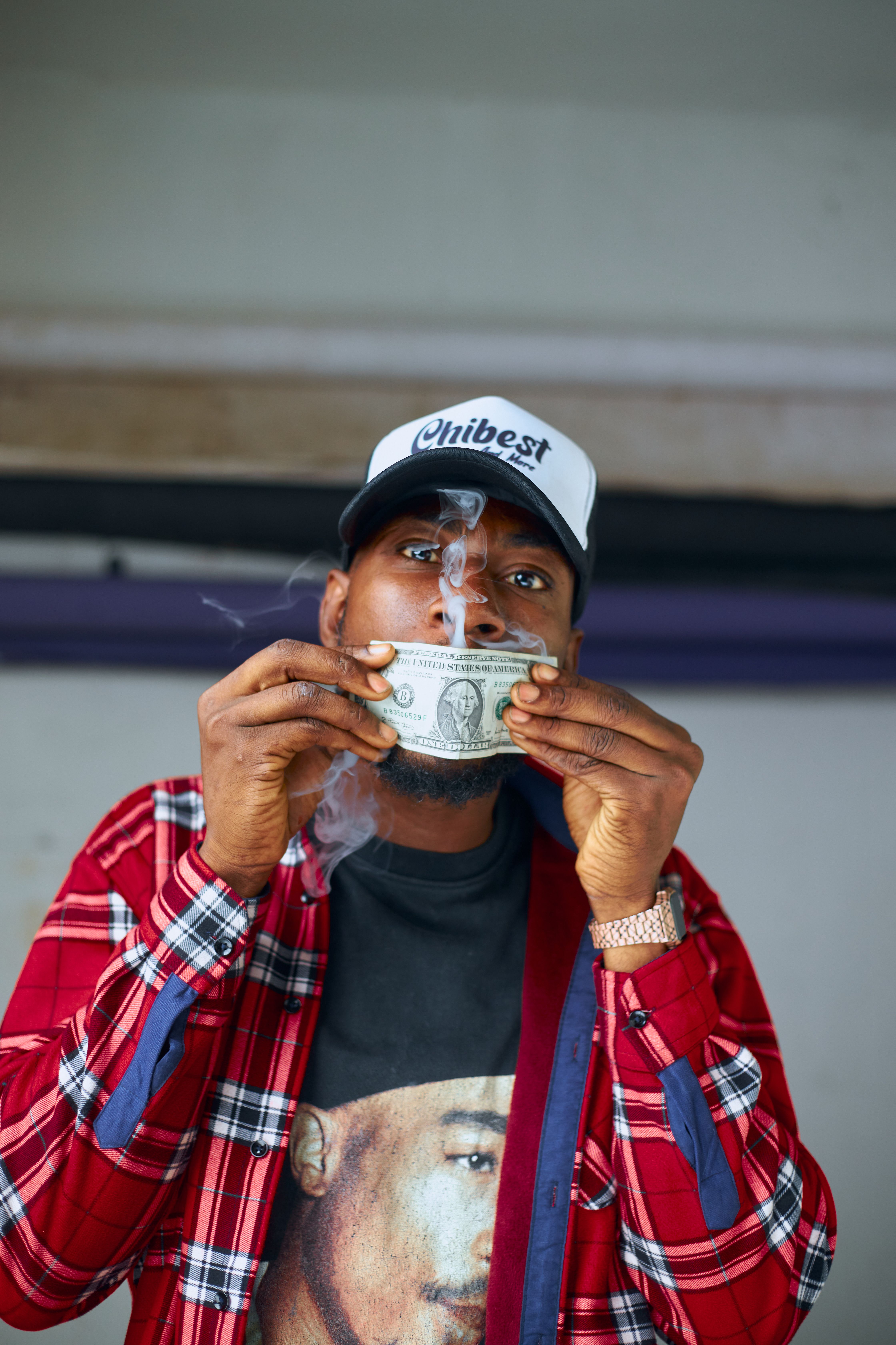 Young man smokes through dollar bill in urban setting wearing a red plaid shirt and cap.