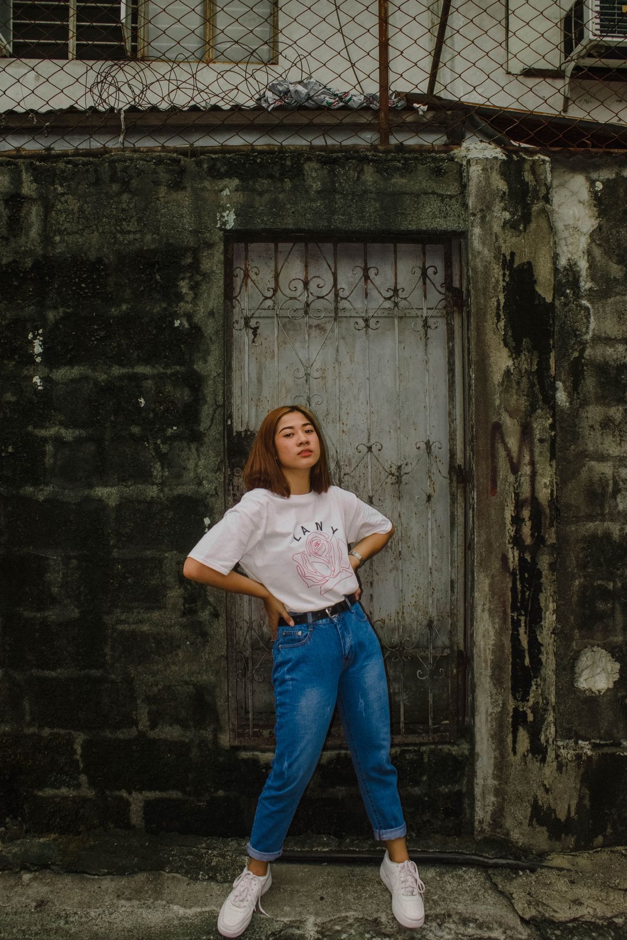 Young woman in a white graphic tee, blue jeans, and white sneakers against a wall