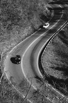 Two cars navigate a winding road through a forest in a black and white photograph.