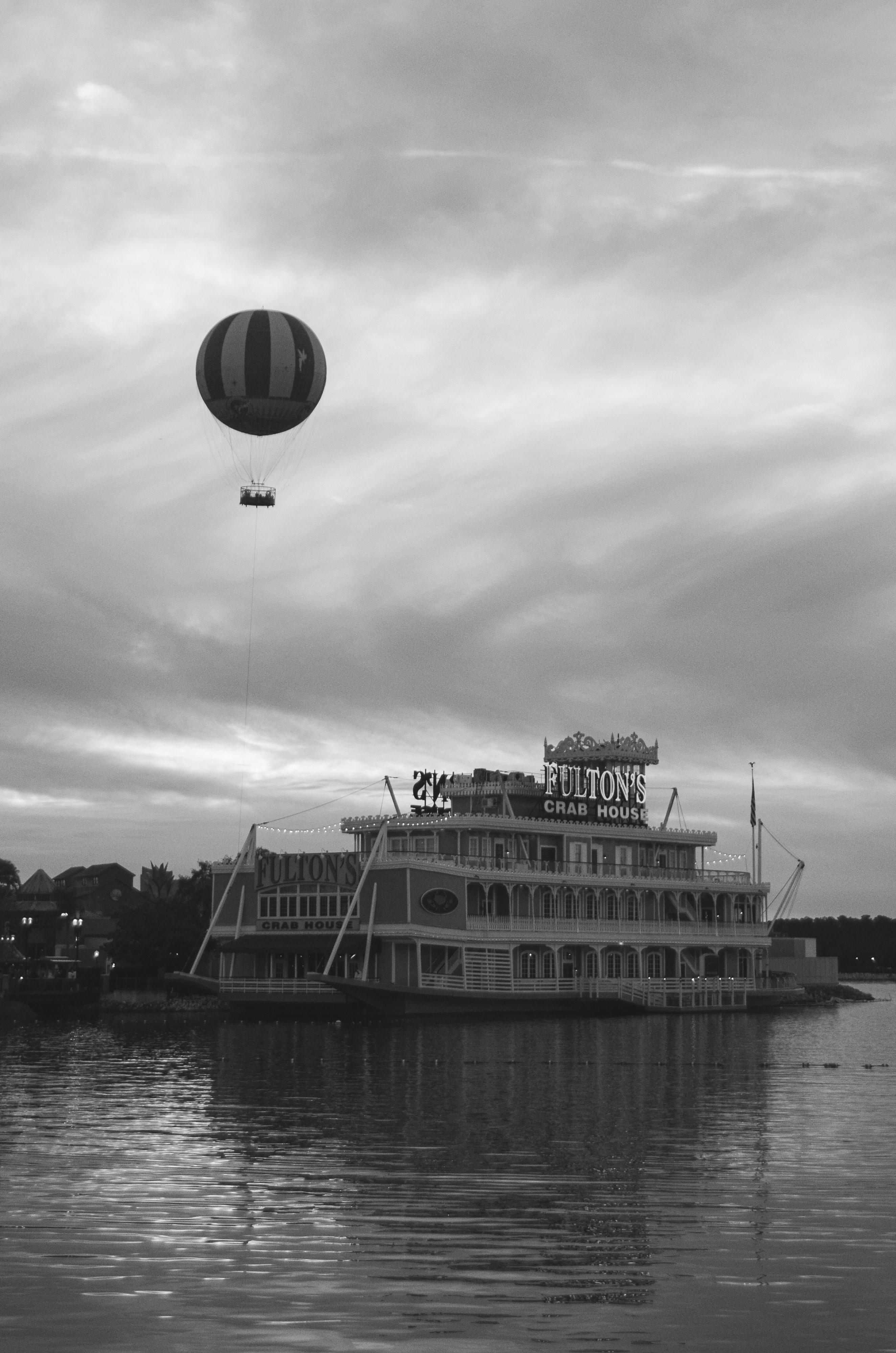 A striking black and white photo of a hot air balloon floating above Fulton's Crab House on a river.
