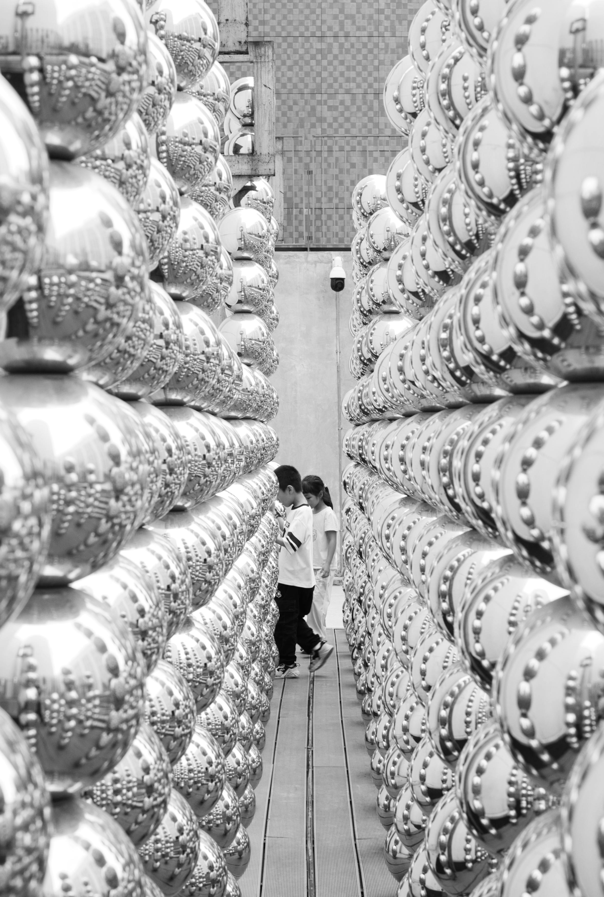 Black and white abstract view of people walking among rows of metallic spheres.