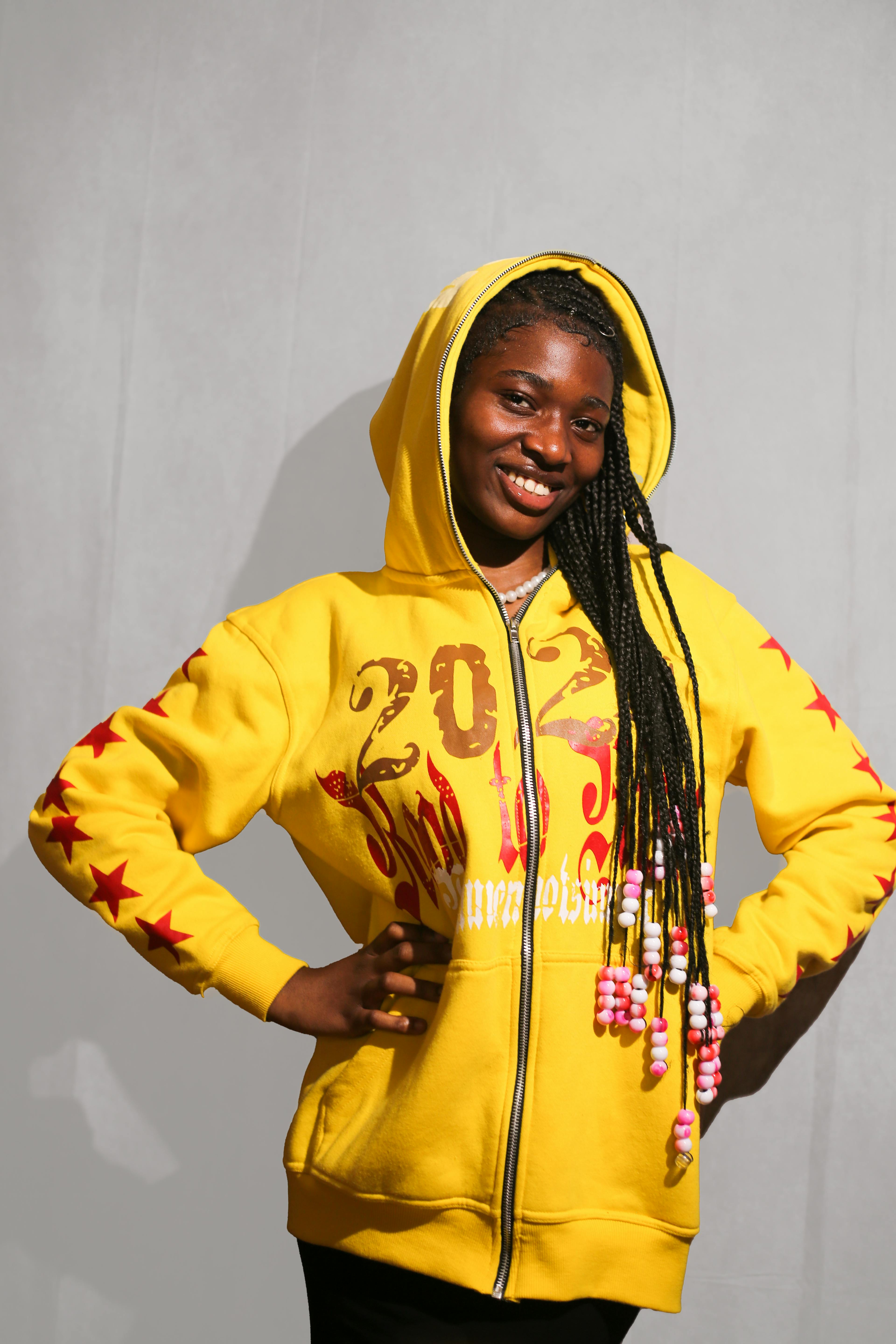 Confident woman in a yellow hoodie with 2023 design against a gray background.