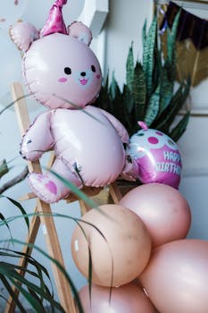Cute pink teddy bear balloon with birthday balloons and tropical plants, creating a festive atmosphere.