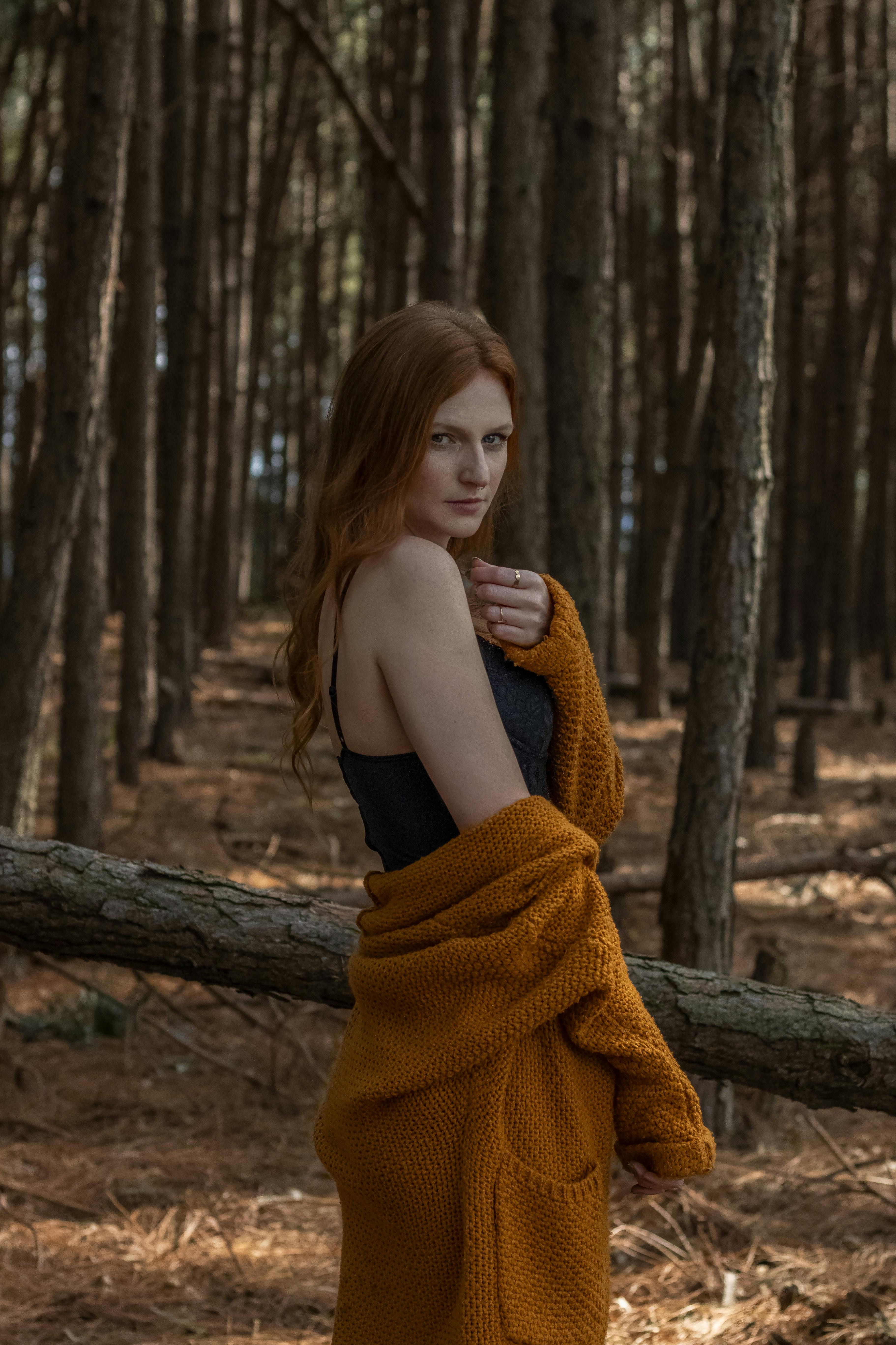 A woman with red hair wrapped in an orange blanket stands in a serene forest setting.