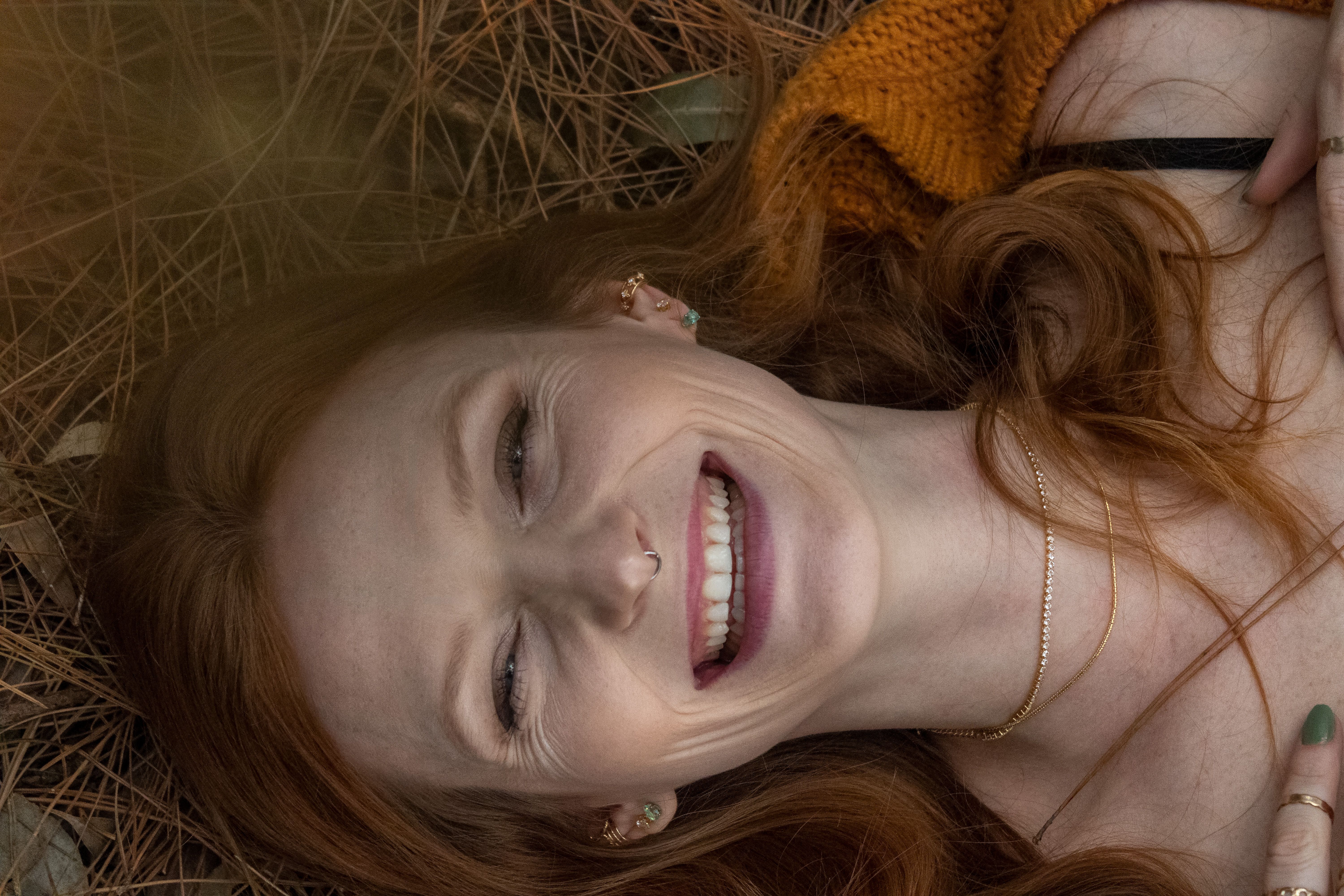 Joyful woman with red hair smiling while lying on pine needles outdoors.