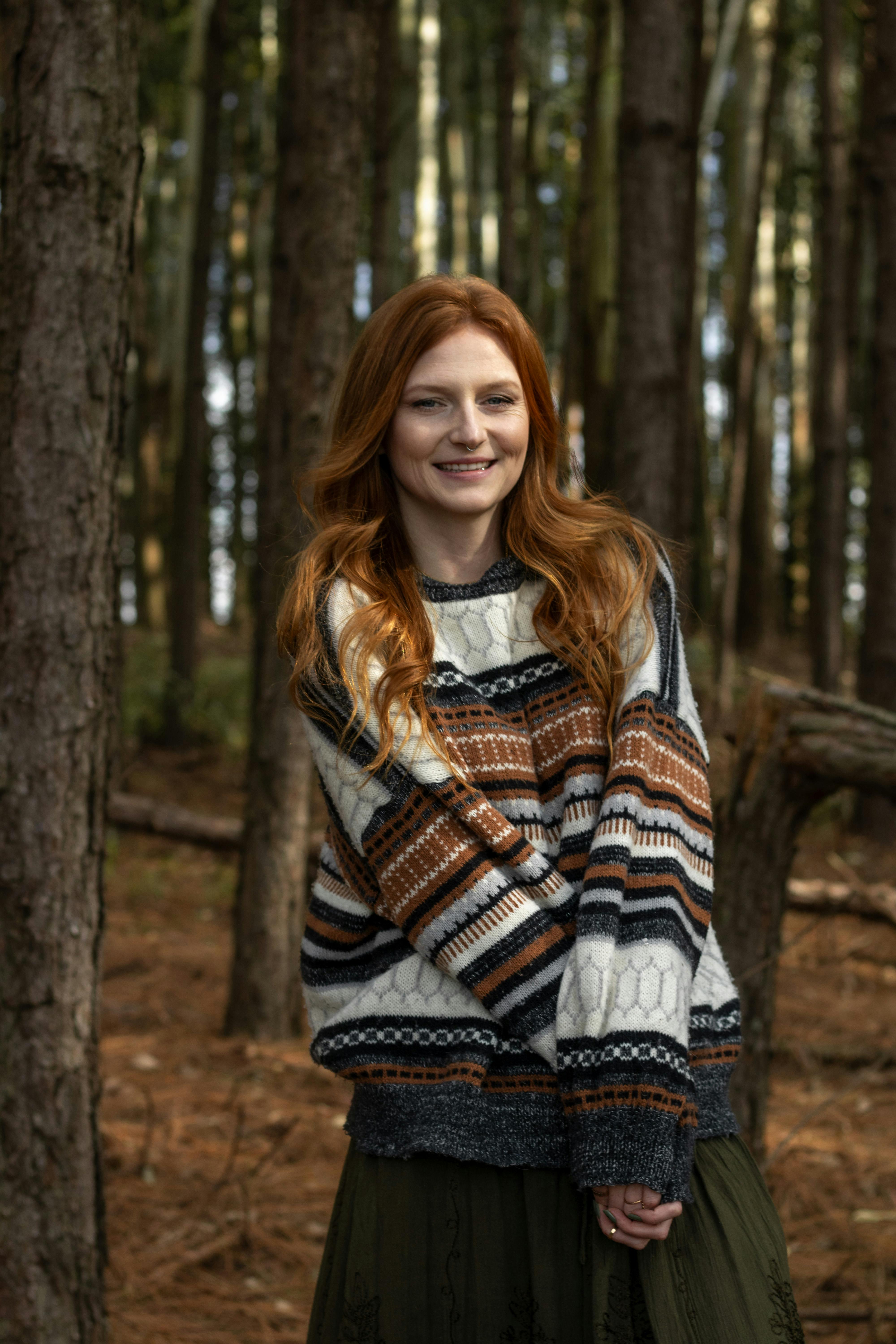 Smiling woman wearing a patterned sweater standing amidst tall trees in autumn.