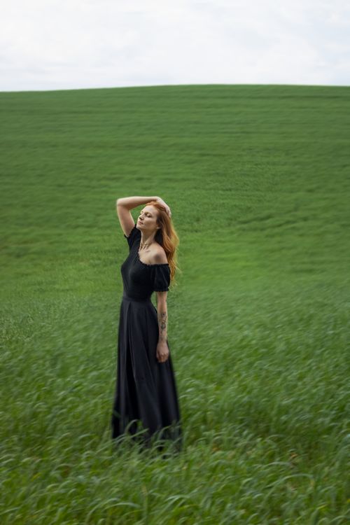Free Serene portrait of a woman in a black dress standing in a lush green field outdoors. Stock Photo
