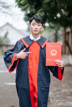 Young man in graduation gown proudly holding diploma outdoors.