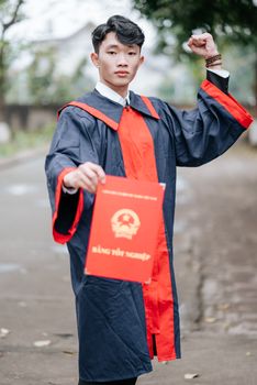 Confident young graduate celebrating with diploma in hand outdoors, wearing a traditional gown.