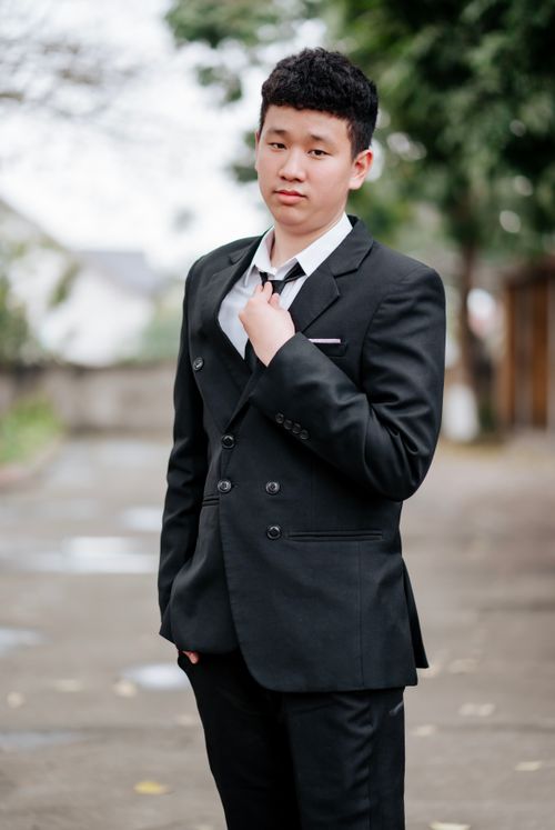 Free A young man confidently adjusting his tie while wearing a black suit outdoors on a cloudy day. Stock Photo