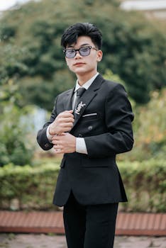 A young man in a sleek black suit poses confidently in an outdoor setting, exuding style and elegance.