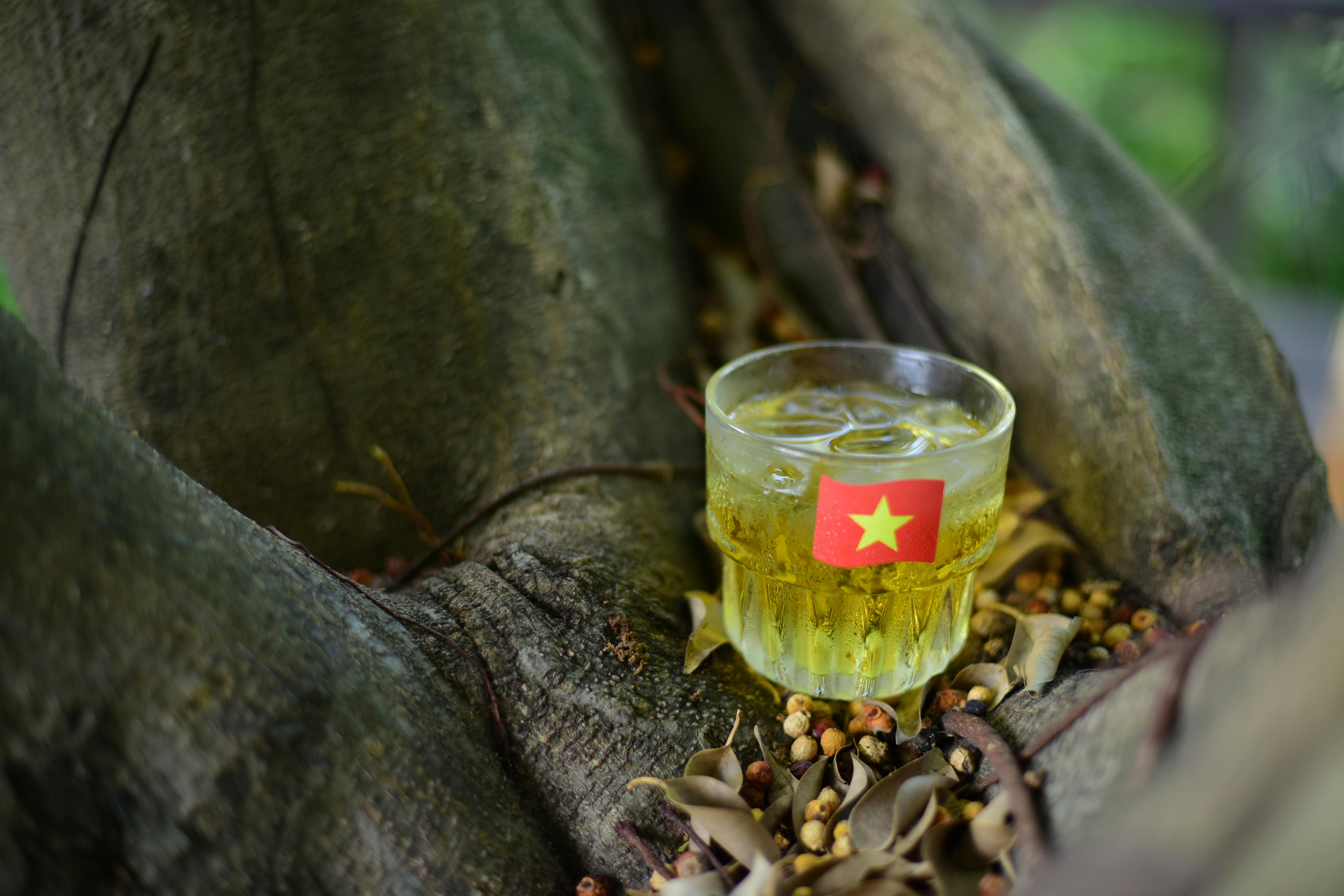 Glass of cold beverage with Vietnamese flag sits on a tree trunk outdoors, surrounded by dry leaves.