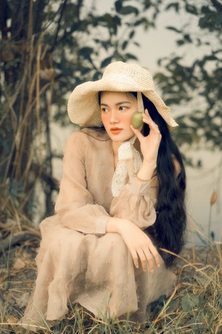 Elegant woman in a white dress holding fruit, sitting in nature wearing a sun hat.