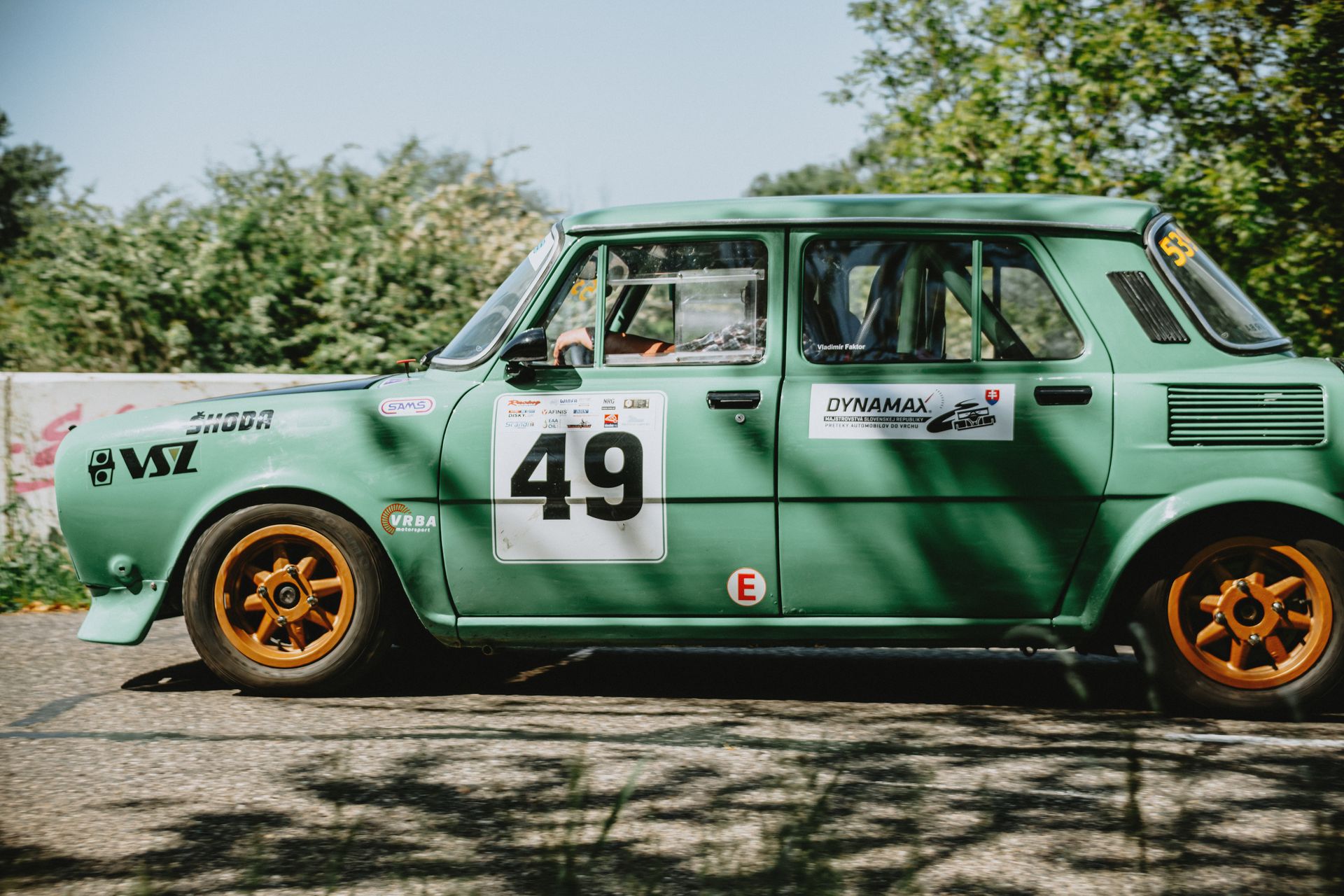 Green vintage Škoda car with number 49 racing on track outdoors surrounded by greenery.