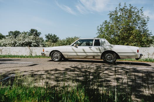 A vintage white classic car driving along a sunny road surrounded by lush greenery.