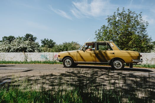 Yellow vintage car driving on a sunny countryside road with lush greenery.