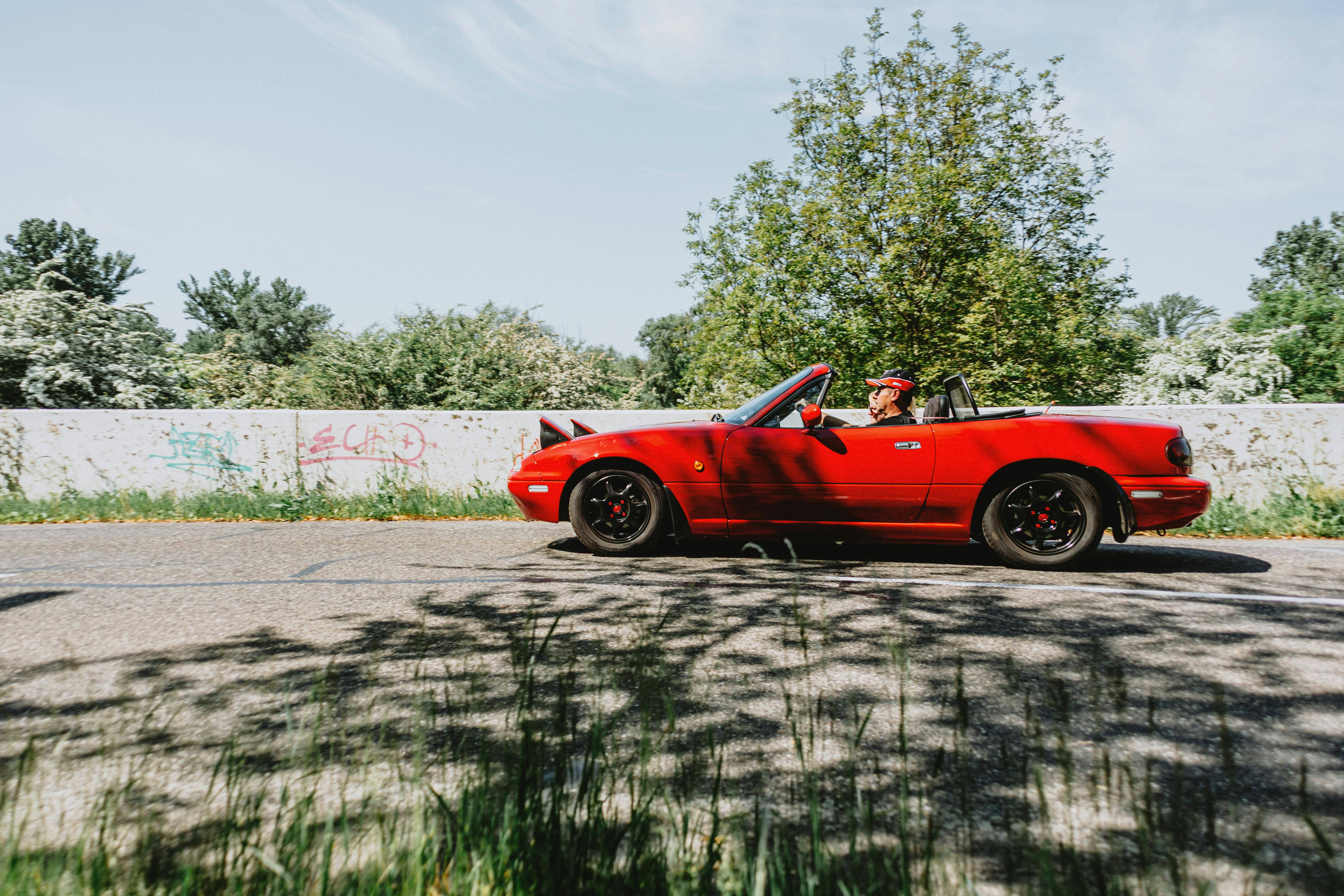 A stylish red convertible car with the roof down, driven by a person on a scenic outdoor road.