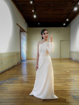 An elegant woman in a white gown poses confidently in a stylish indoor setting with wooden floors.