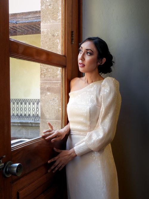 Free A fashionable woman in an elegant white dress stands by a sunlit doorway, exuding beauty and grace. Stock Photo