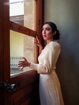 Elegant woman in a white dress gazing thoughtfully by a sunlit window indoors.