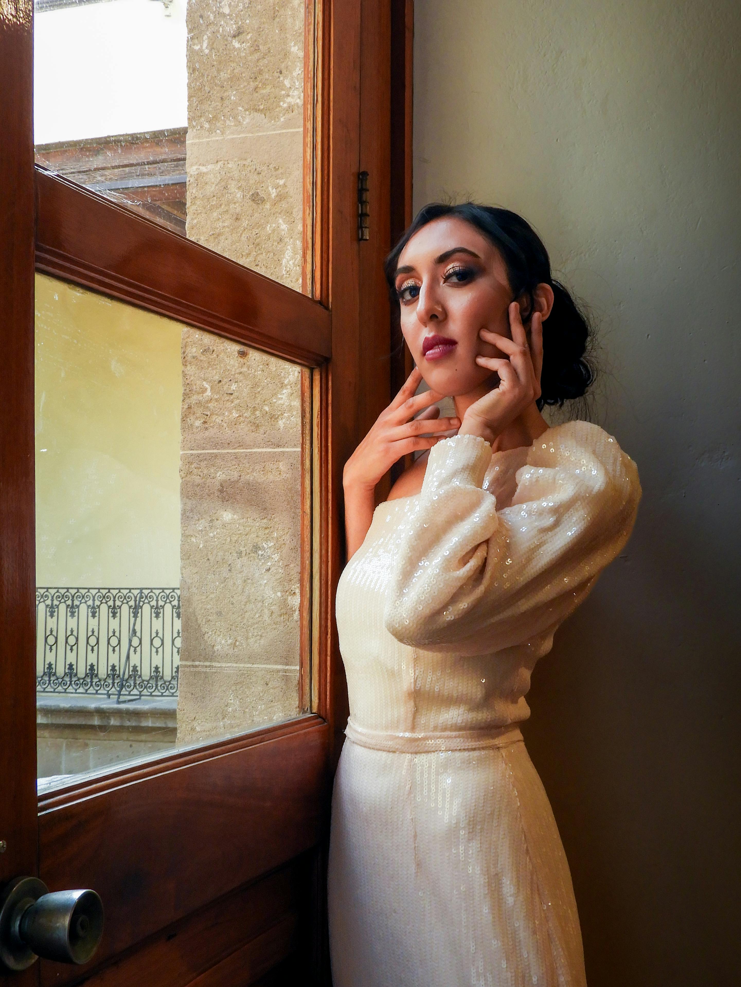 Sophisticated woman in a glamorous dress poses elegantly by a window, showcasing timeless fashion.