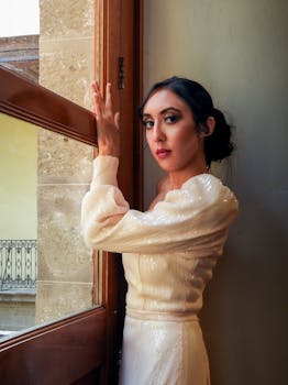 Woman in elegant attire posing by a window, exuding grace and style indoors.