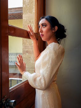 A woman in an elegant white gown gazes thoughtfully out a window, creating a serene and sophisticated scene.