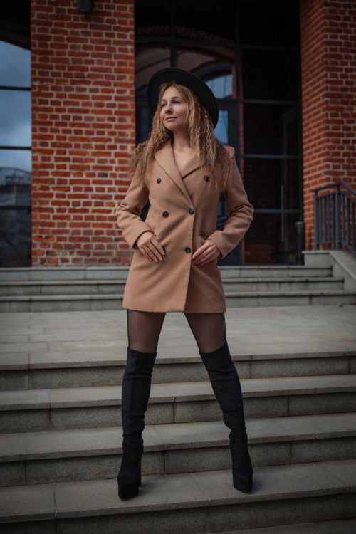 Free Stylish woman in coat and boots standing confidently on city steps against brick wall. Stock Photo