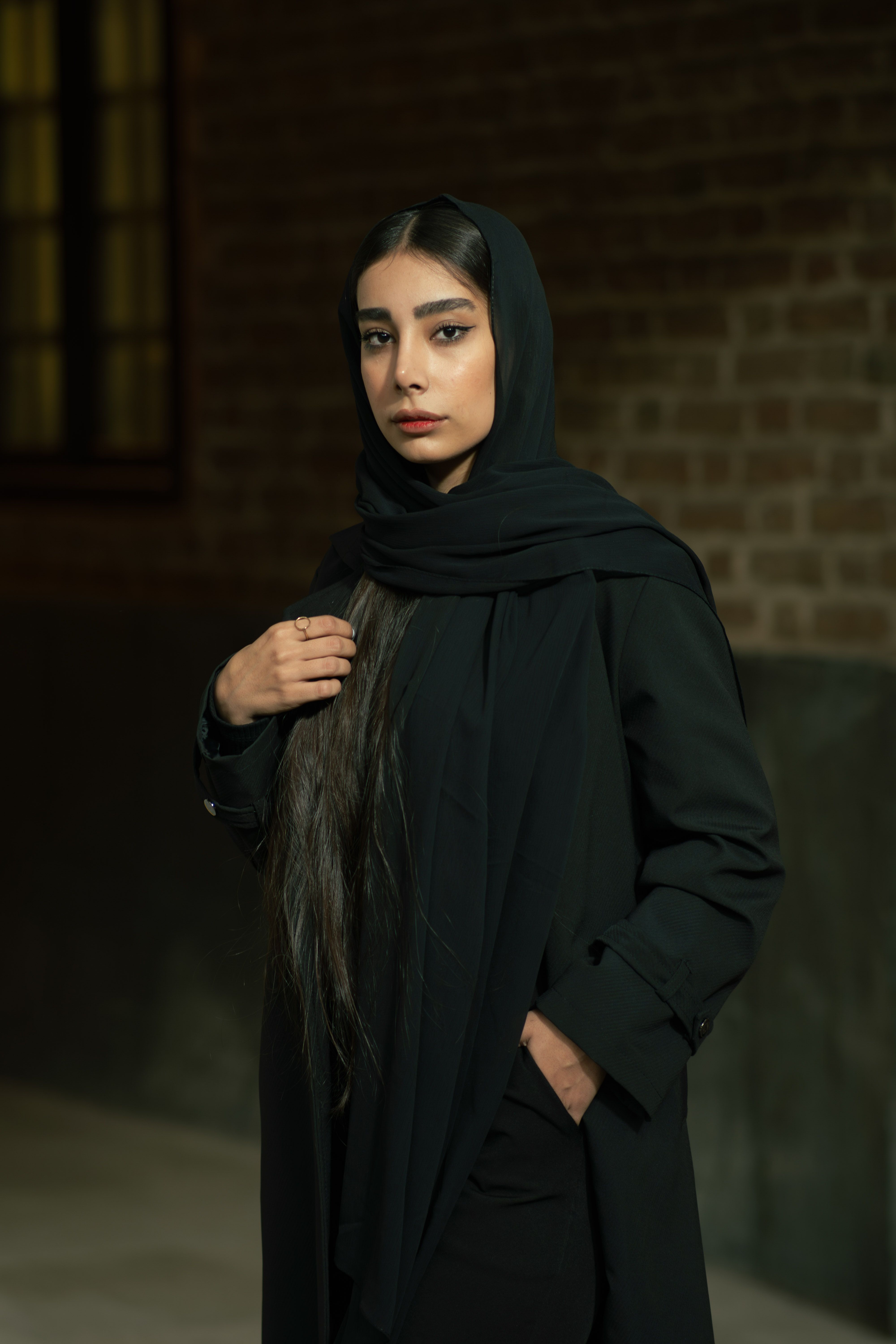 Elegant portrait of a young woman in a black hijab against a brick wall in Tehran.