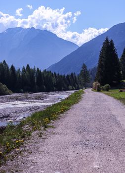 A tranquil dirt path runs alongside a river with a mountain backdrop under a clear blue sky.