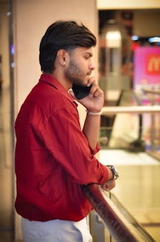 A young man in a red shirt talks on the phone inside a shopping mall.