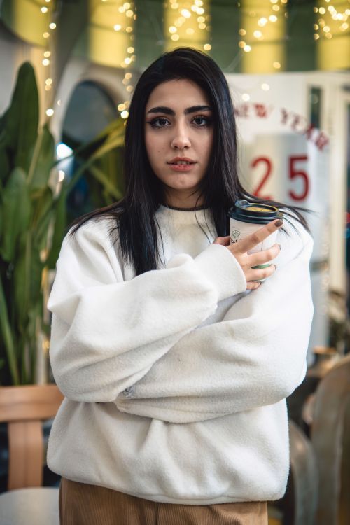 Free Young woman in white sweater holding coffee, captured in a festive Tehran setting. Stock Photo