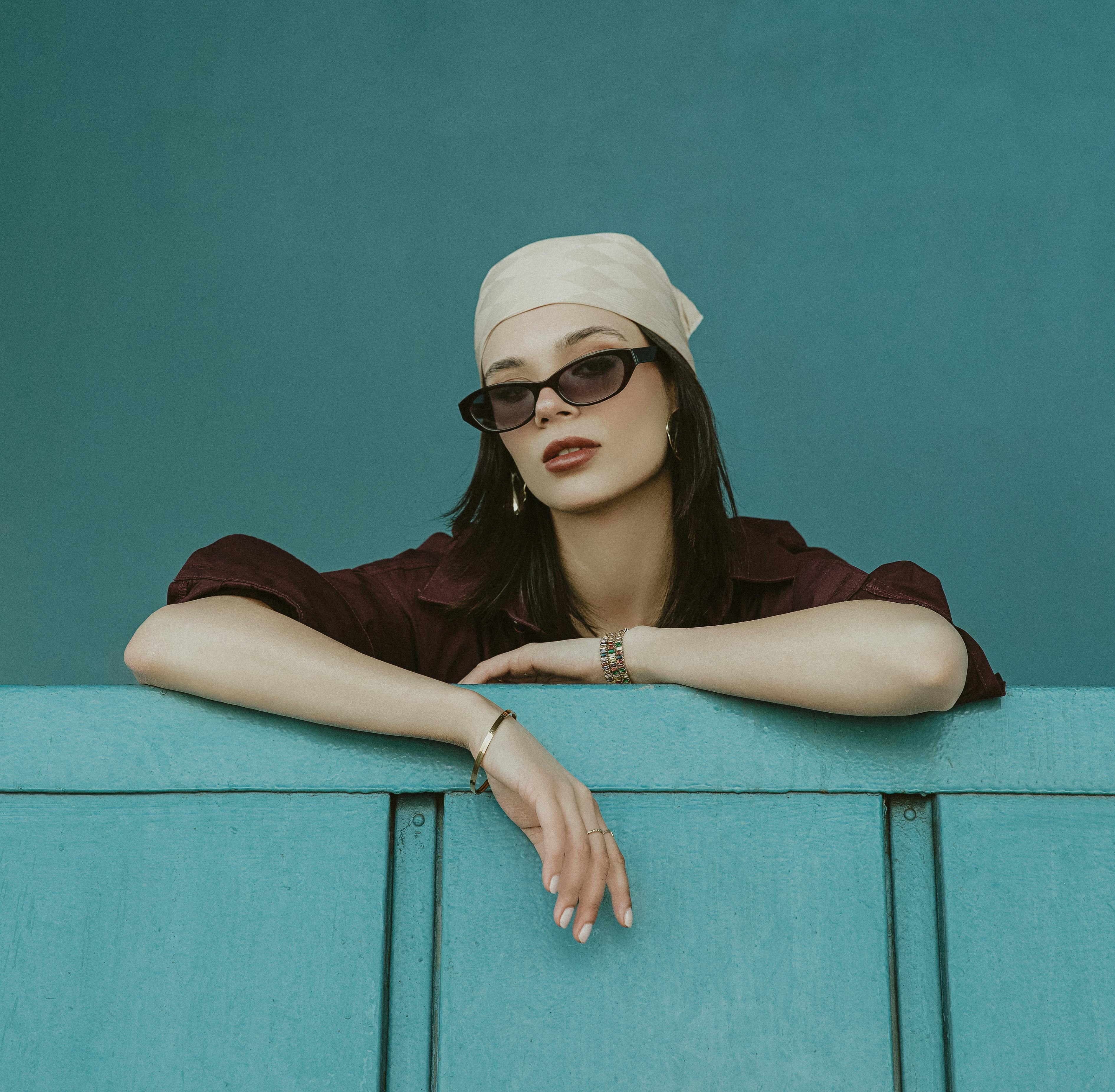 Chic model posing in İstanbul with sunglasses and headscarf against blue backdrop.