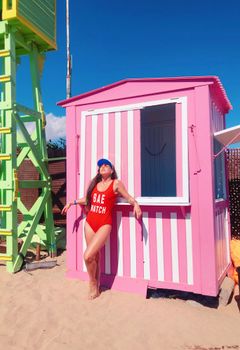 Woman in red swimsuit enjoying a sunny beach day by a pink striped cabana.