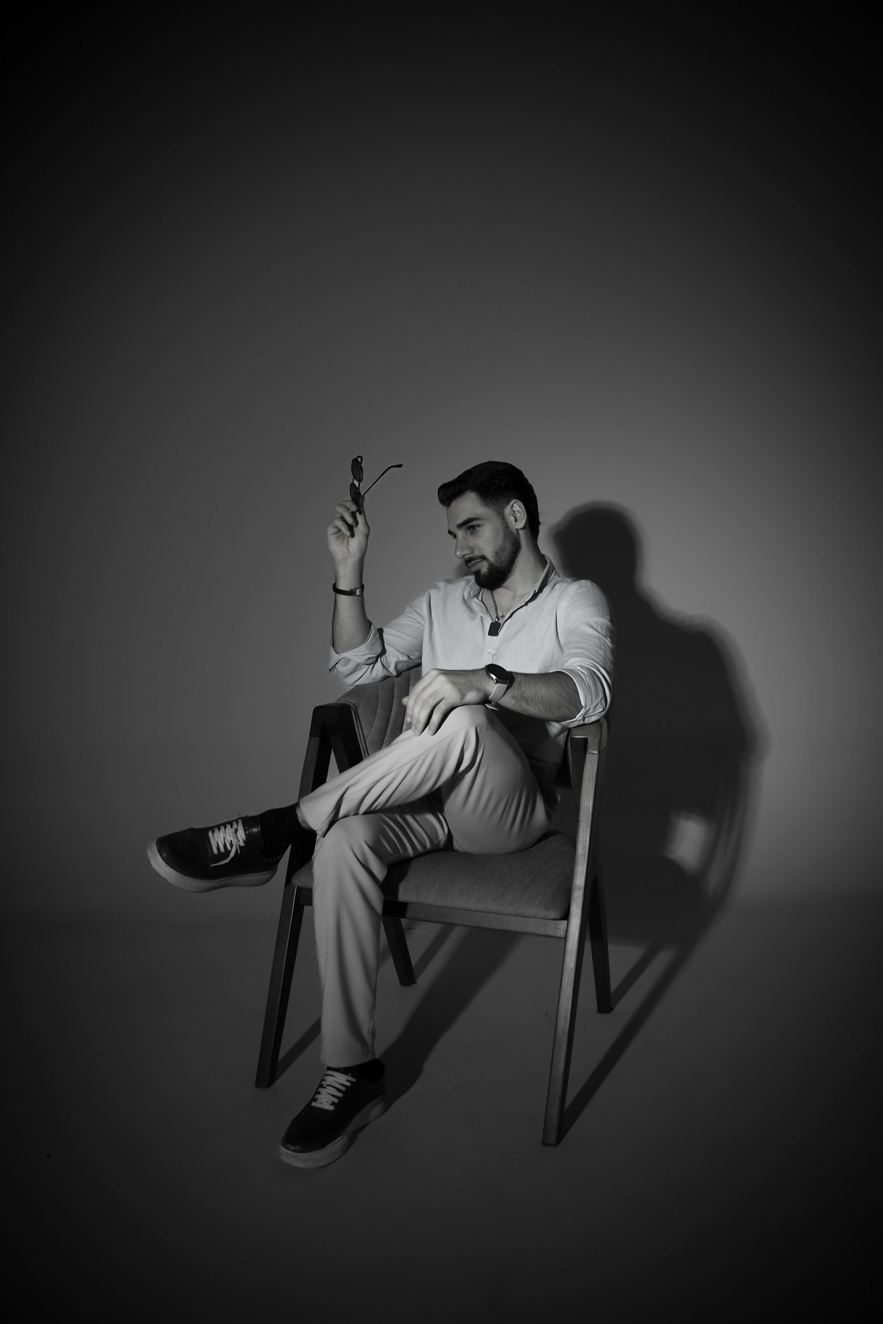 Zadarmo Štýlový muž v monochromatickom štýle sediaci na stoličke so slnečnými okuliarmi v ruke s dramatickým osvetlením. Fotka z fotobanky