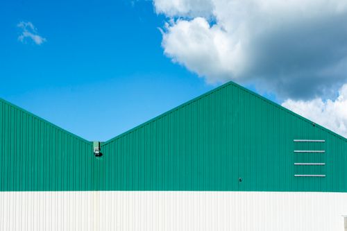 Free Modern green warehouse facade with angled roof under clear blue sky and fluffy clouds. Stock Photo