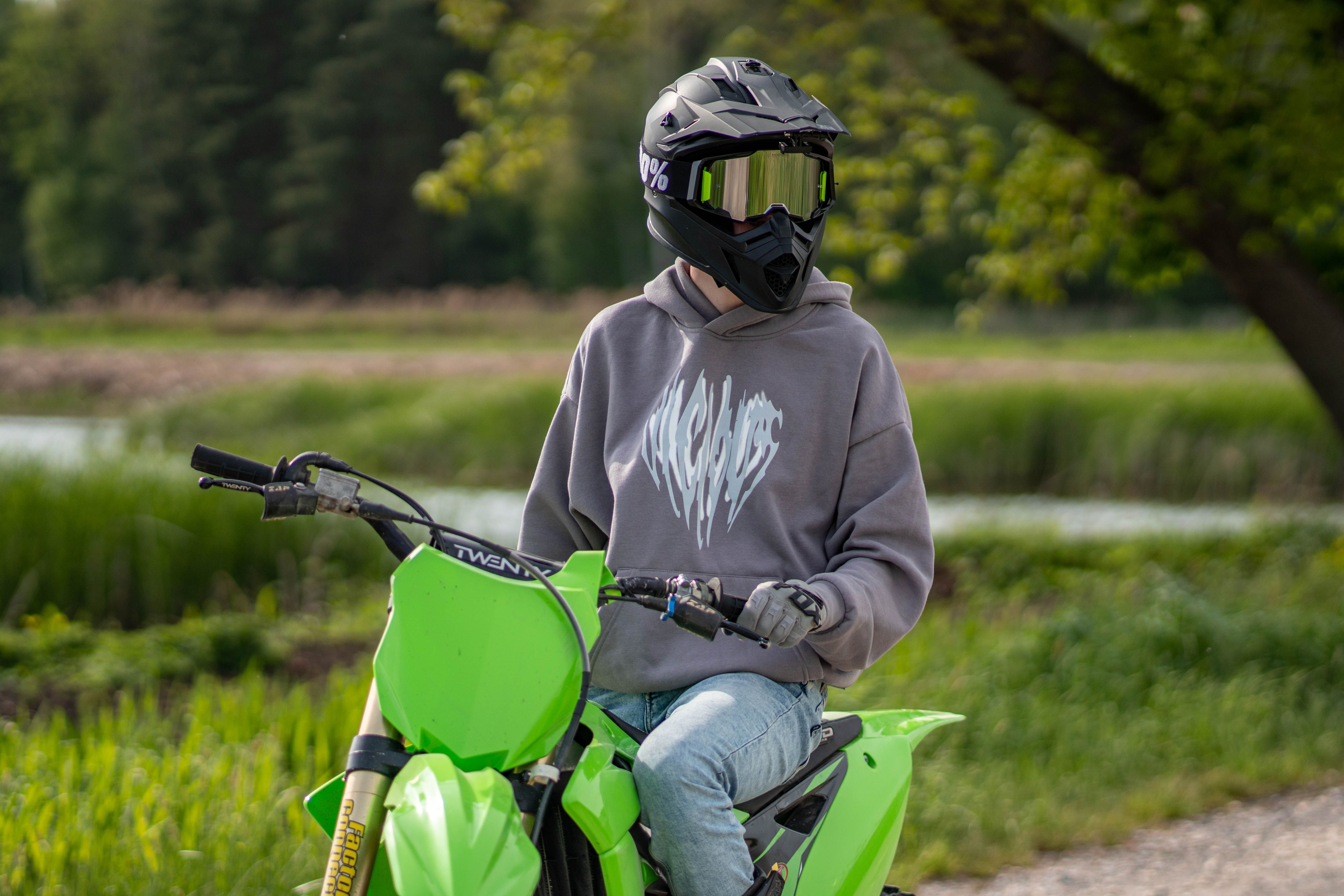 Person on a green dirt bike wearing a helmet and hoodie in a rural outdoor setting.