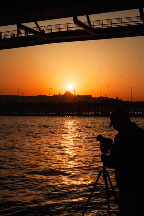 Free Silhouette of a photographer capturing a vibrant sunset over the water from under a bridge. Stock Photo