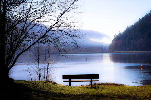 Serene landscape of a lake with an empty bench and misty hills in the background.