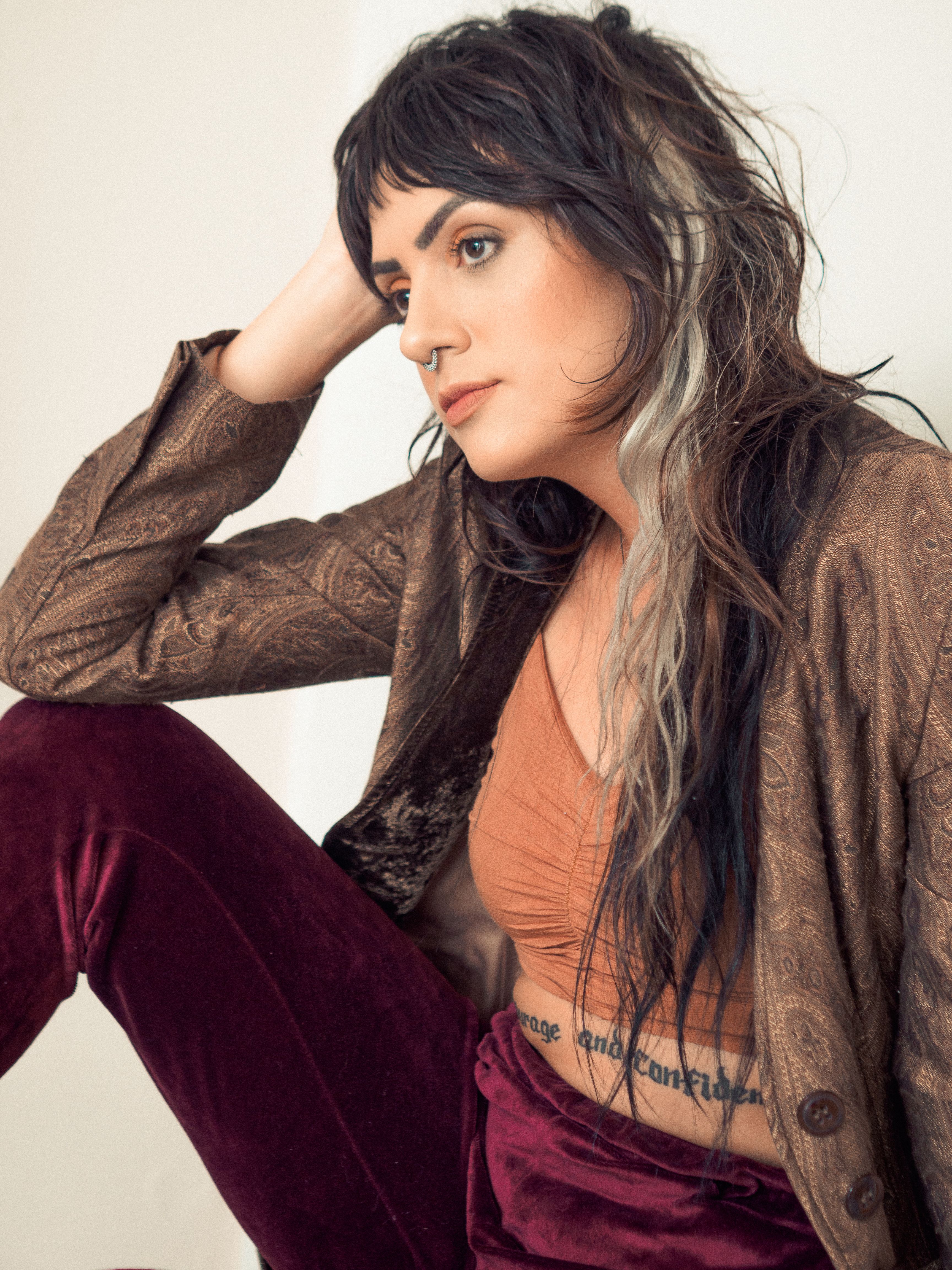 A fashionable young woman with long hair and tattoos poses thoughtfully indoors in vintage attire.
