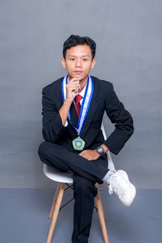 Confident young man in formal attire with a medal posing seated on a chair.