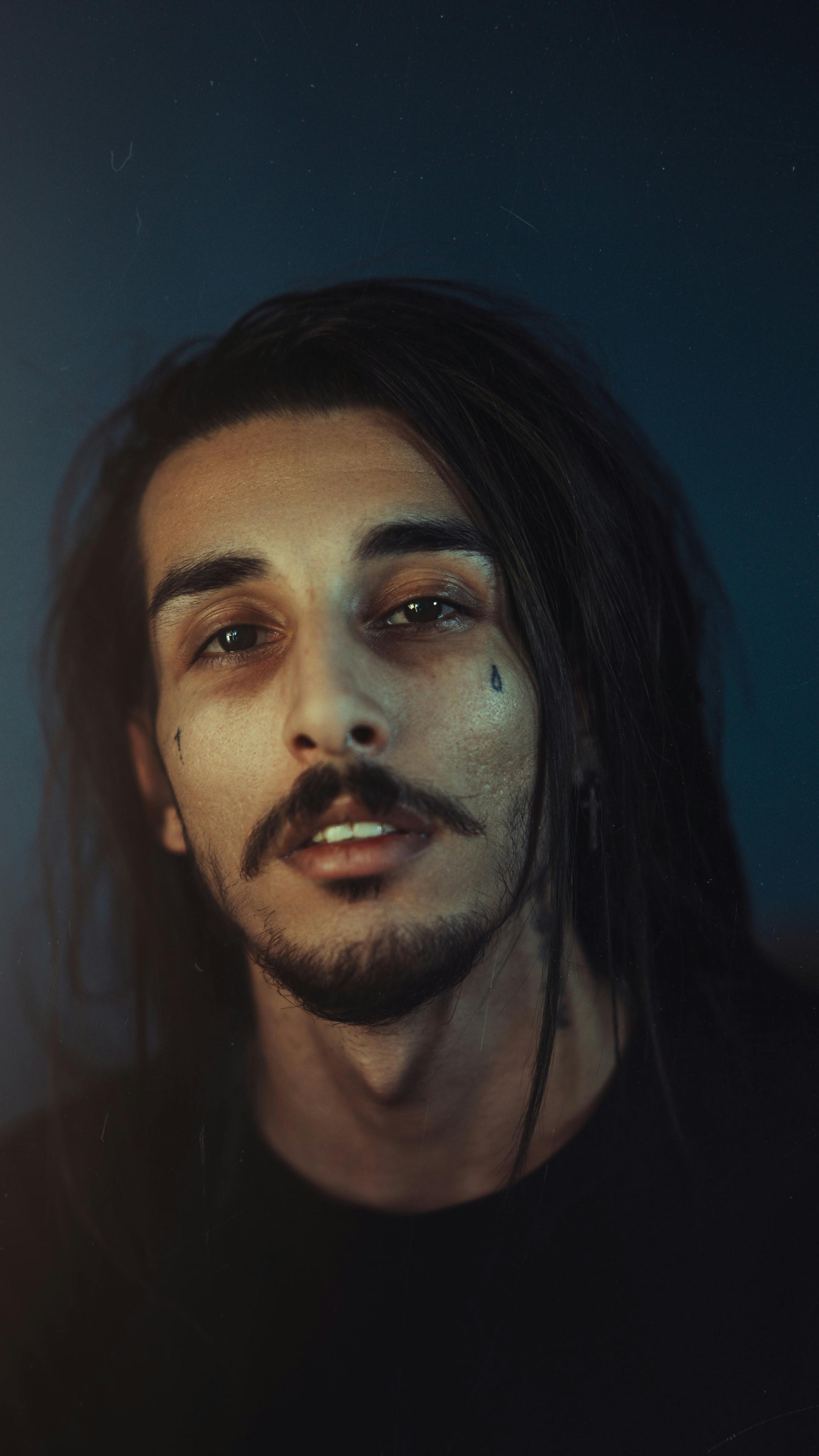 A dramatic, moody portrait of a young man with long hair and teardrop tattoos against a dark background.