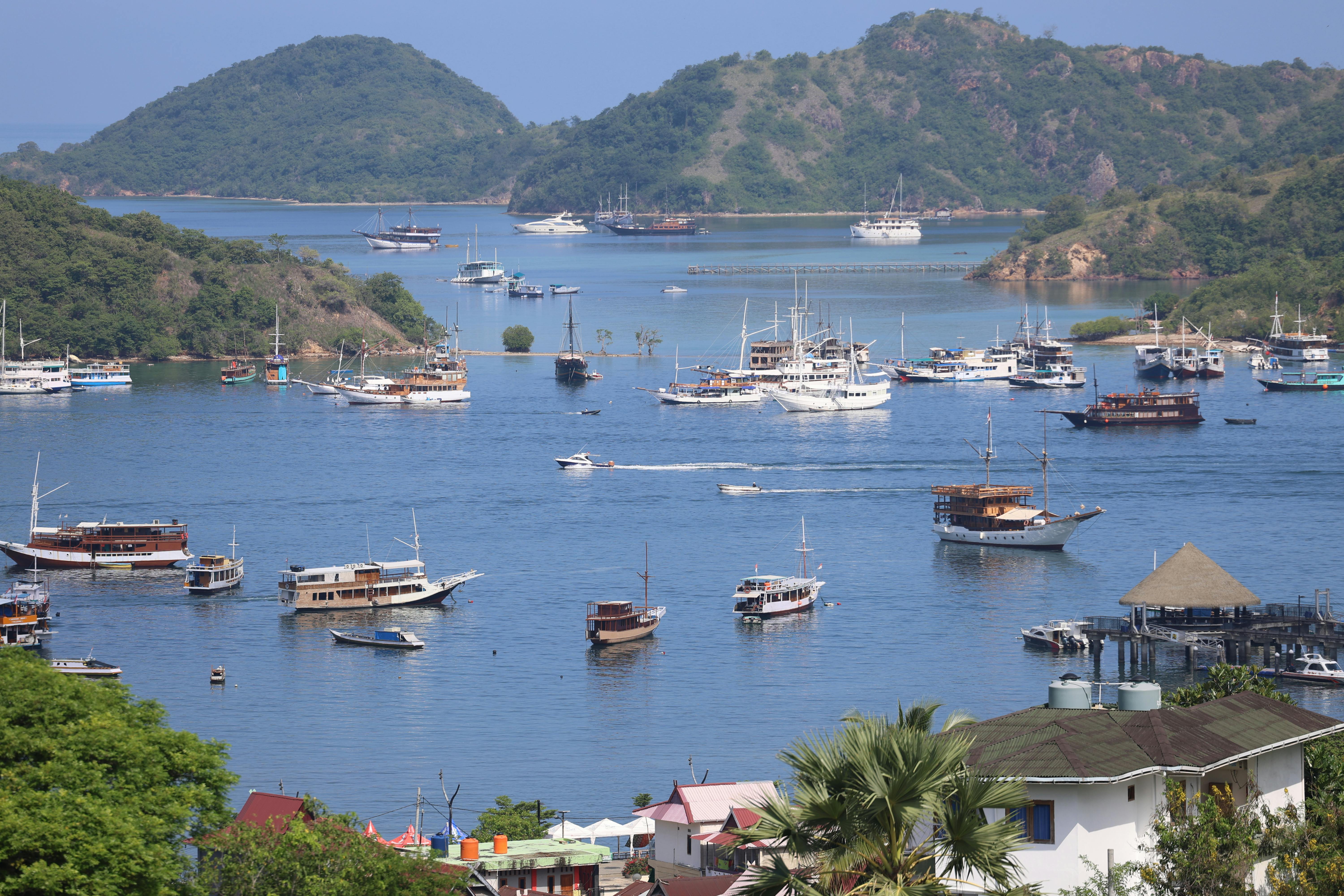 Meanwhile, Labuan Bajo port comes into view with dozens of liveaboard boats and yachts anchored in a calm bay, framed by green hills and a small waterfront pier.