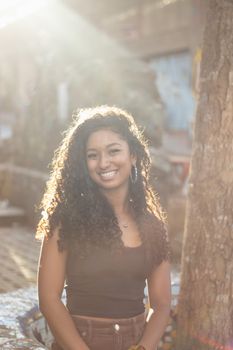 A woman with curly hair smiles warmly in outdoor sunlight.