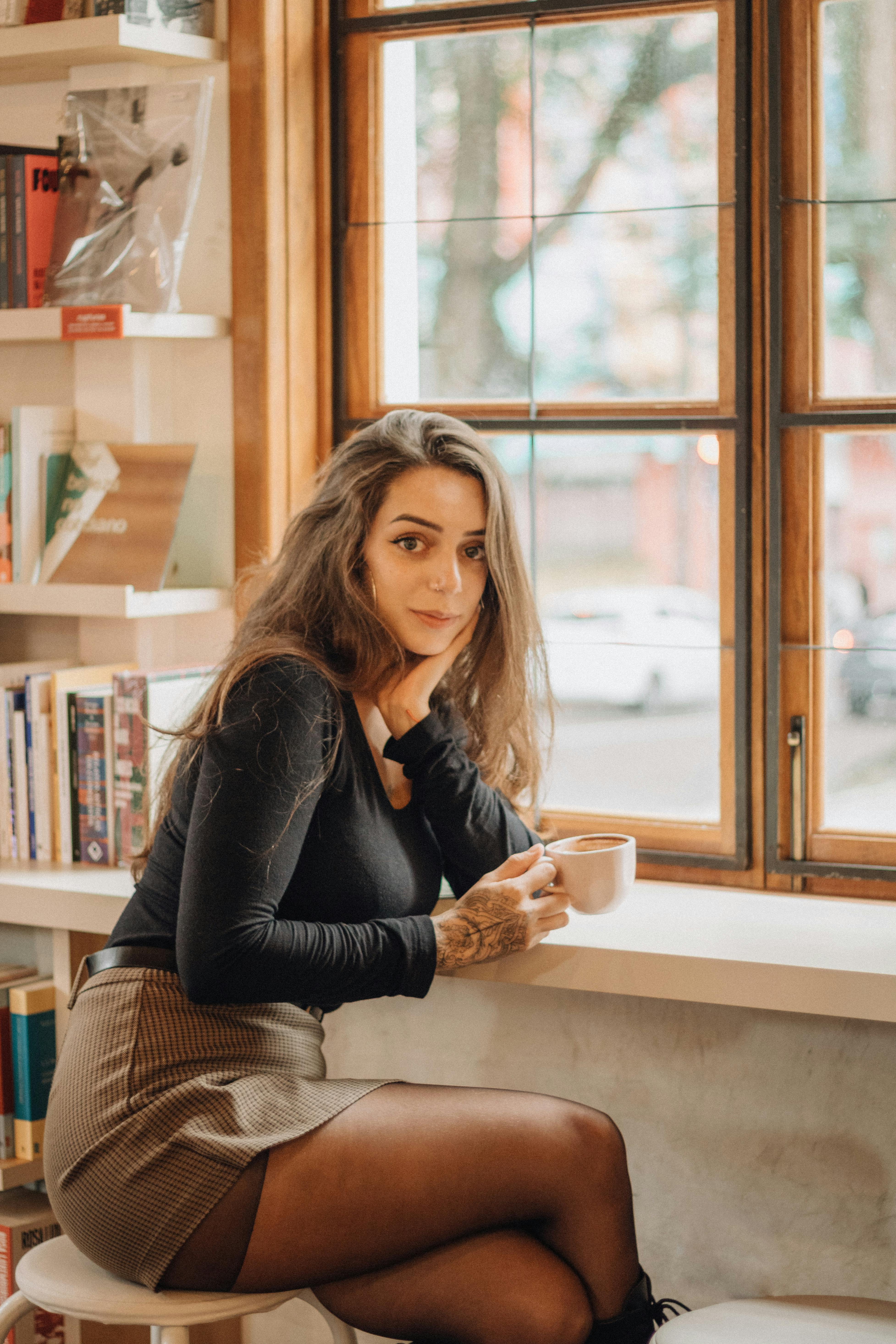 De franc Dona jove gaudint d'un cafè a la vora d'una finestra en un cafè acollidor, envoltada de llibres. Foto d'estoc