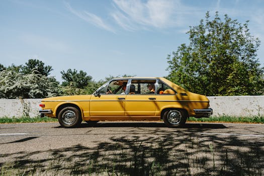 A classic yellow car drives along a scenic road surrounded by lush greenery and clear blue skies.