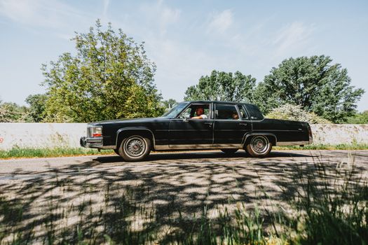 Vintage black sedan cruising on a scenic road on a sunny day, surrounded by lush greenery.