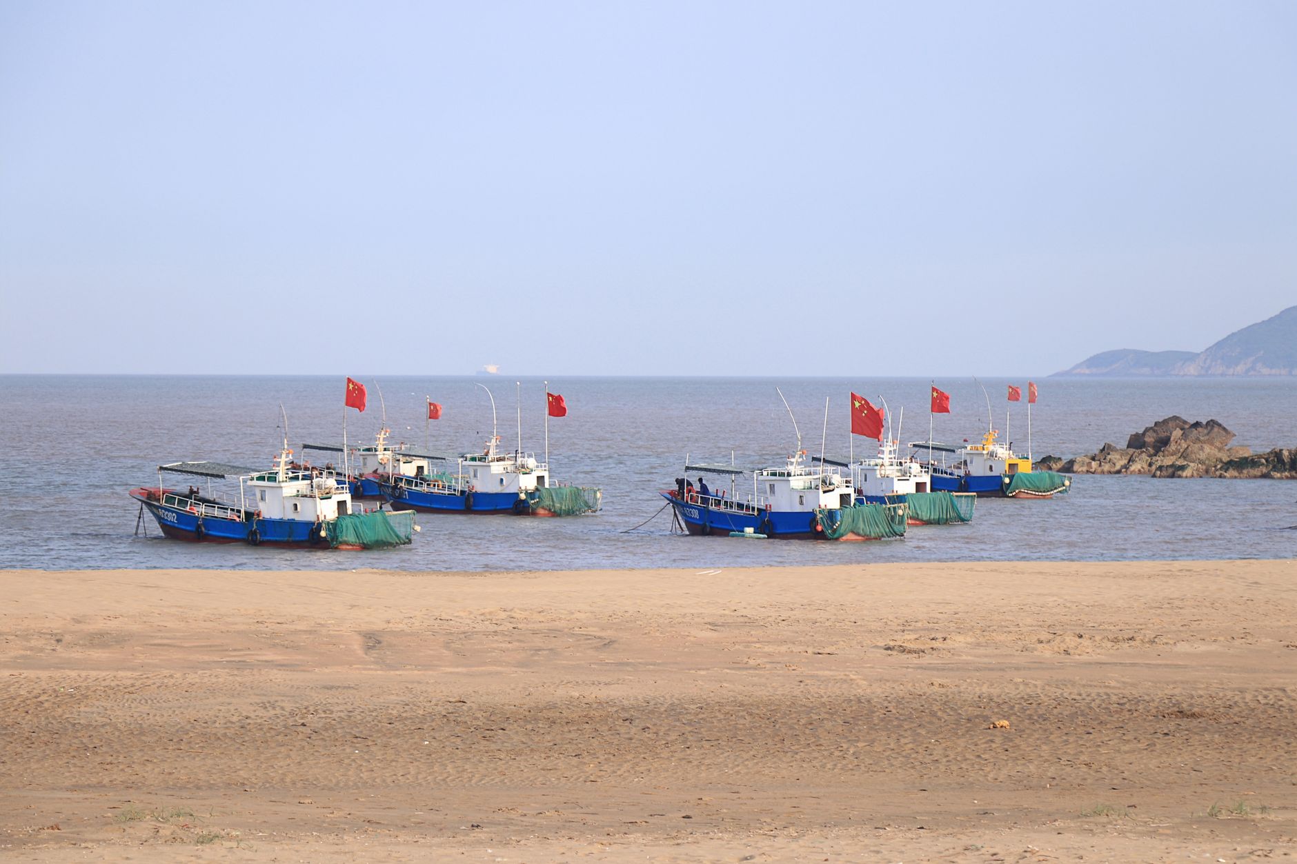 Fishing boats with red flags anchored on the sandy beach with a clear blue sky backdrop.